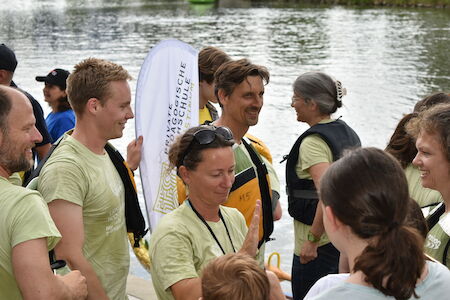 Eine Gruppe von Menschen in Schwimmwesten versammelt sich lächelnd und unterhaltend am Flussufer.