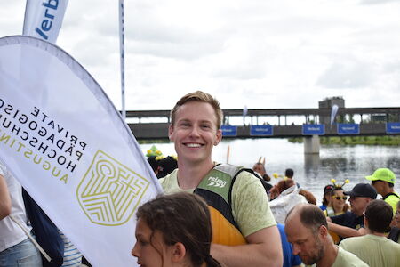 Lächelnder Mann mit einem Paddel bei einer Veranstaltung am Flussufer mit Menschenmenge und Fahnen.