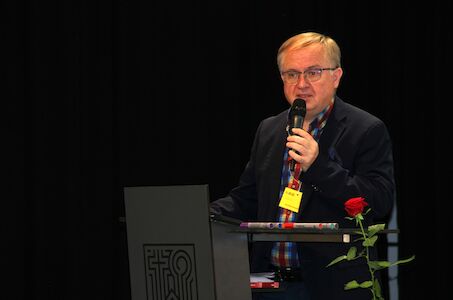 Mann spricht in ein Mikrofon an einem Podium mit einer roten Rose an der Seite.