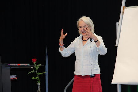 Ältere Frau gestikuliert beim Sprechen, neben einem Flipchart und einer einzelnen roten Rose.