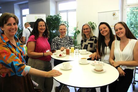 Sechs lächelnde Frauen stehen mit Getränken und Kuchen um einen Tisch herum.
