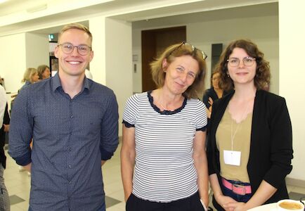 Drei Personen stehen lächelnd bei einer Veranstaltung oder Konferenz in geschlossenen Räumen zusammen.