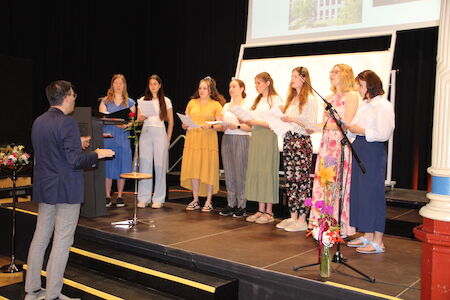 Eine Gruppe von Frauen singt auf der Bühne, während ein Mann sie dirigiert.