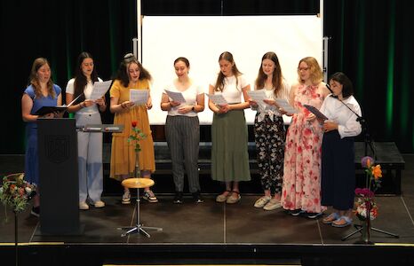 Neun Frauen stehen auf einer Bühne und singen gemeinsam, sie halten Blätter Papier in den Händen.