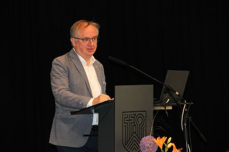 Ein Mann im grauen Blazer spricht an einem Podium mit einem Laptop und Blumen in der Nähe.