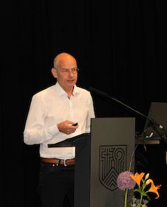 Mann im weißen Hemd spricht an einem Podium mit Blumen im Vordergrund.