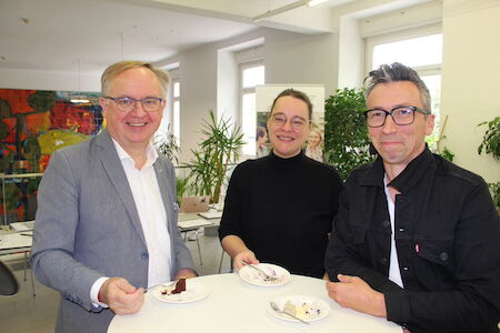 Drei Personen lächeln und essen Kuchen an einem Tisch in einem hellen Innenbereich.