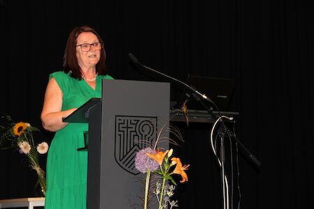 Frau im grünen Kleid spricht an einem Podium mit Blumen und schwarzem Hintergrund.