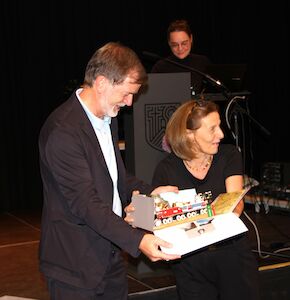 Zwei Menschen lächeln und halten eine Kiste mit bunten Spielzeugfahrzeugen auf einer Bühne.