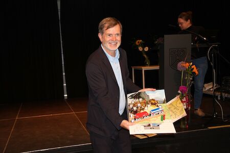 Ein lächelnder Mann im Anzug hält ein farbenfrohes Kunstwerk auf einer Bühne mit Blumen in der Nähe.