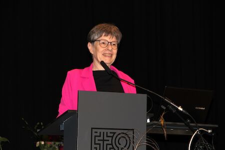 Eine Frau in einer rosa Jacke spricht an einem Podium mit einem Mikrofon.