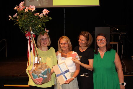 Vier lächelnde Frauen, eine hält einen Blumenkorb und eine andere ein verpacktes Geschenk.