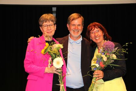 Drei lächelnde Menschen, zwei davon mit Blumensträußen in der Hand, stehen vor einem schwarzen Vorhang.