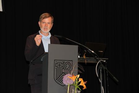Mann spricht an einem Podium mit Blumen und einem Laptop vor einem schwarzen Hintergrund.