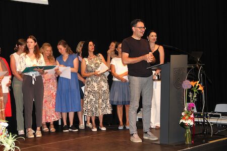 Ein Mann spricht an einem Podium; mehrere Frauen stehen mit Papieren in der Hand hinter ihm.