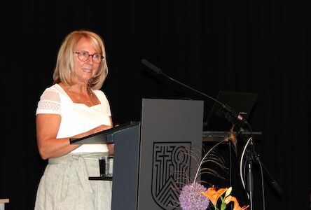 Frau mit Brille spricht an einem Podium, Blumen und Laptop in der Nähe.