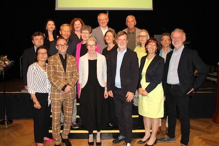 Eine Gruppe von fünfzehn Erwachsenen posiert gemeinsam im Innenbereich und lächelt in die Kamera.