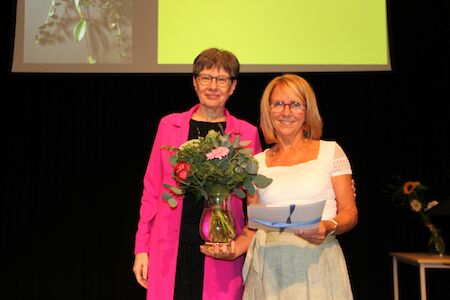 Zwei Frauen stehen zusammen, eine hält eine Vase mit Blumen und einen Ordner.