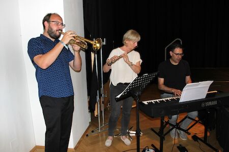 Drei Musiker spielen gemeinsam Trompete, Flöte und Keyboard im Innenbereich vor einer Bühne.