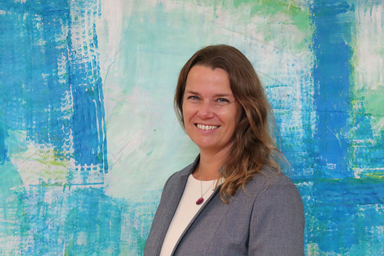 Frau mit langen braunen Haaren, die vor einem abstrakten blauen Hintergrund lächelt.