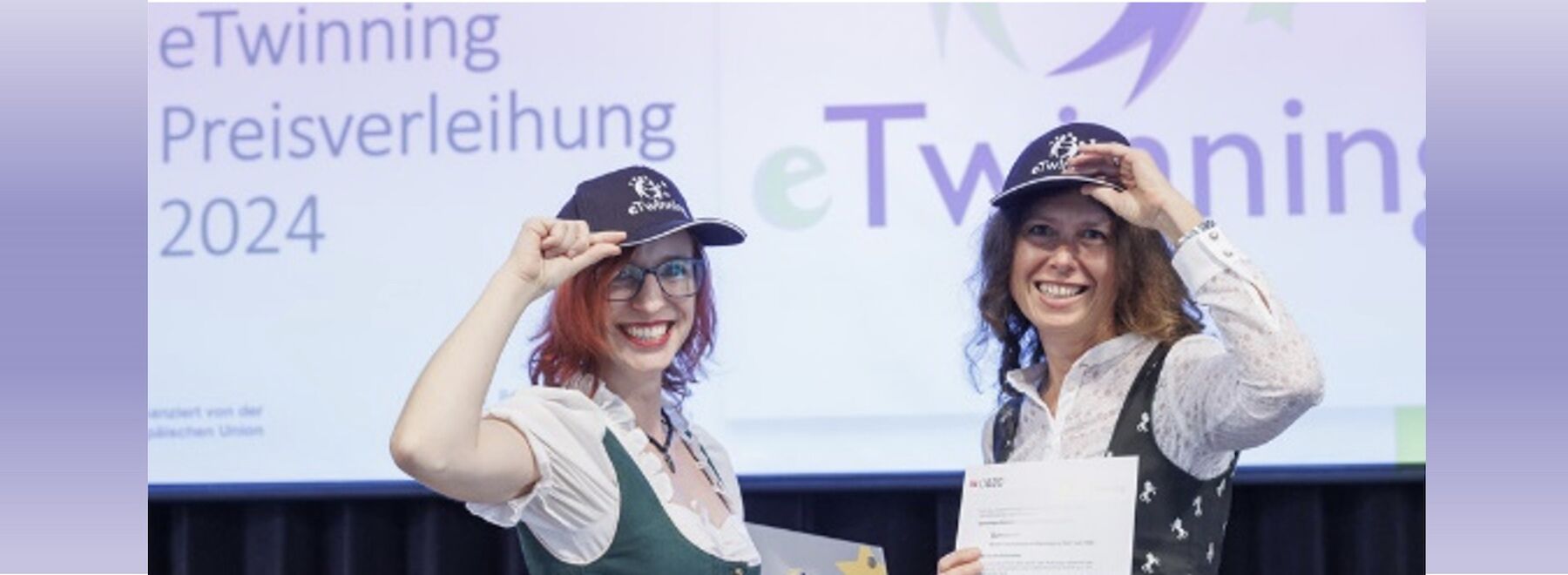 Zwei Frauen mit eTwinning-Hüten lächeln und halten Zertifikate bei einer Preisverleihung.