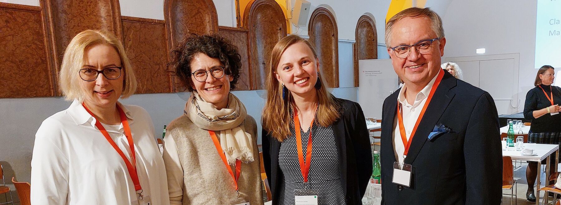Vier lächelnde Erwachsene stehen mit roten Schlüsselbändern im Innenbereich einer Konferenzveranstaltung.