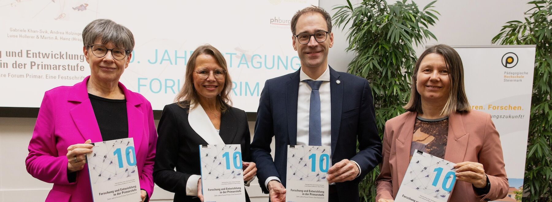 Vier Personen in Geschäftskleidung halten bei einer formellen Veranstaltung Broschüren hoch.
