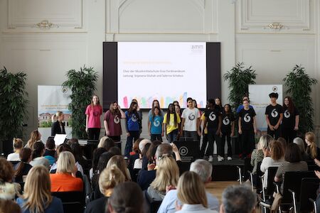 Eine Gruppe von Menschen steht auf der Bühne und singt für ein Publikum in einem großen Saal.