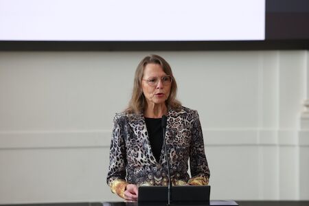 Frau im gemusterten Blazer spricht an einem Podium in einem hellen Raum.