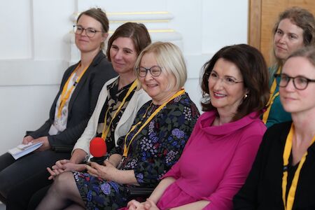 Sechs Frauen sitzen und lächeln bei einer Veranstaltung, alle tragen gelbe Schlüsselbänder.