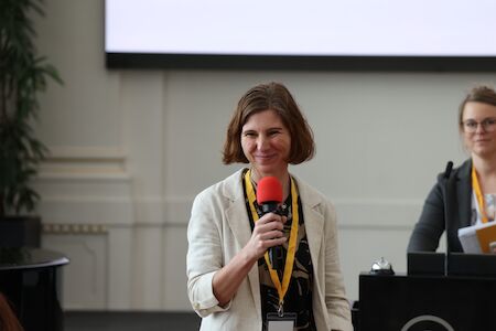 Frau lächelt und spricht bei einer Veranstaltung in einem geschlossenen Raum in ein rotes Mikrofon.