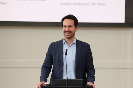 Mann im Anzug lächelt und spricht an einem Podium mit Mikrofon.