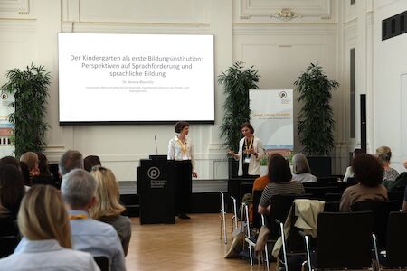Zwei Frauen präsentieren vor einem Publikum in einem Konferenzraum mit einer großen Leinwand.