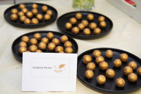 Vier schwarze Teller mit goldenen runden Bonbons auf einem Tisch, beschriftet mit „Goldene Perlen“.
