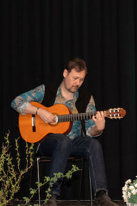Mann spielt im Sitzen Akustikgitarre, im Vordergrund Pflanzen.