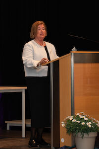 Eine Frau steht an einem Podium und spricht. Vor dem Podium stehen Blumen.