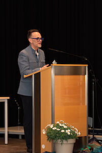 Person mit Brille spricht an einem Podium, davor steht eine Topfpflanze.