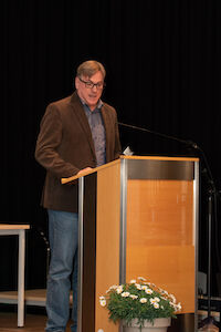 Ein Mann mit Brille spricht an einem Podium, davor eine Pflanze, vor einem dunklen Hintergrund.
