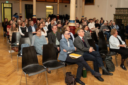 Das Publikum sitzt in Reihen und verfolgt aufmerksam eine Präsentation in einem großen Raum.