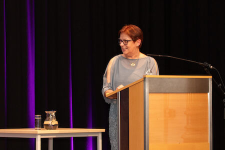 Frau spricht an einem Podium mit einem Mikrofon, einem Tisch und einem Wasserkrug in der Nähe.