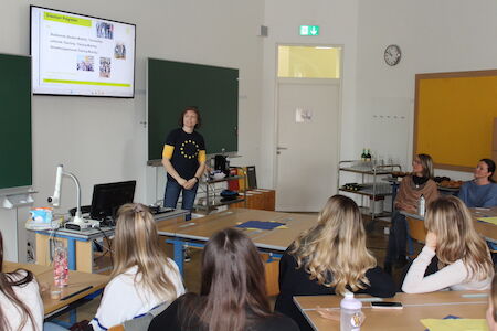 Eine Frau präsentiert einer Gruppe von Schülern in einem Klassenzimmer mit Bildschirmanzeige etwas.