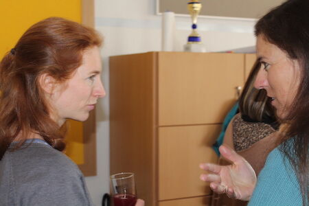 Zwei Frauen unterhalten sich, eine hält ein Glas, im Hintergrund ist ein Schrank zu sehen.