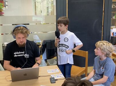 Drei Jungen nehmen an einem Tisch Audio auf, einer hält ein Mikrofon, einer Kopfhörer.