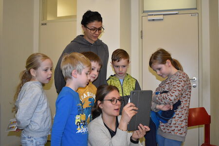 Zwei Erwachsene und fünf Kinder schauen gemeinsam in einem Raum auf ein Tablet.