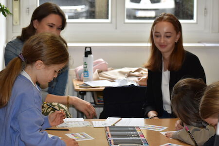 Zwei Frauen und drei Kinder sitzen an einem Tisch und zeichnen und schreiben gemeinsam.