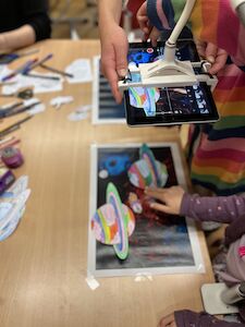 Kinder scannen mit einem Tablet bunte Papierplaneten auf einem Arbeitsblatt zum Thema Weltraum.