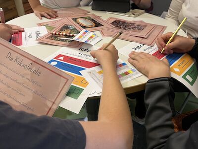 An einem runden Tisch arbeiten Menschen gemeinsam an bunten Arbeitsblättern, eines davon auf Deutsch.