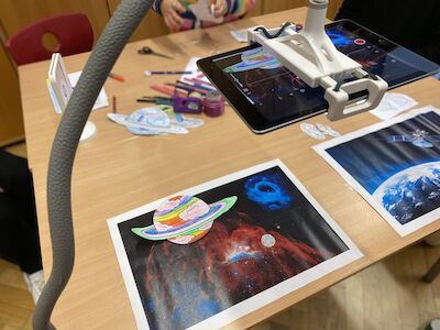 Tablet auf einem Ständer über Kunstdrucken mit Weltraummotiven und Bastelbedarf auf einem Tisch.