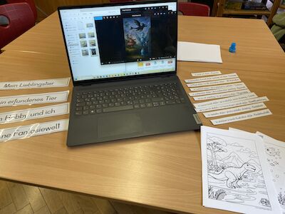 Ein Laptop auf einem Tisch mit deutschen Arbeitsblättern und Dinosaurier-Malvorlagen.