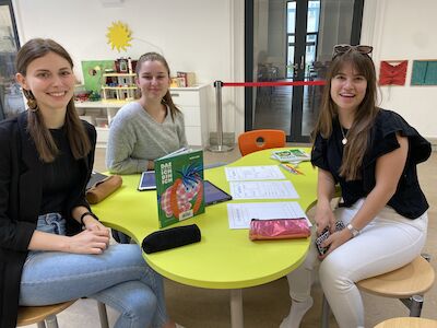 Drei junge Frauen lächeln und sitzen mit Papieren und Büchern um einen gelben Tisch.